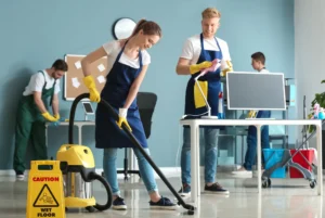 A team of cleaners wearing gloves and aprons tidying an office. One person vacuums the floor while another wipes a computer screen.